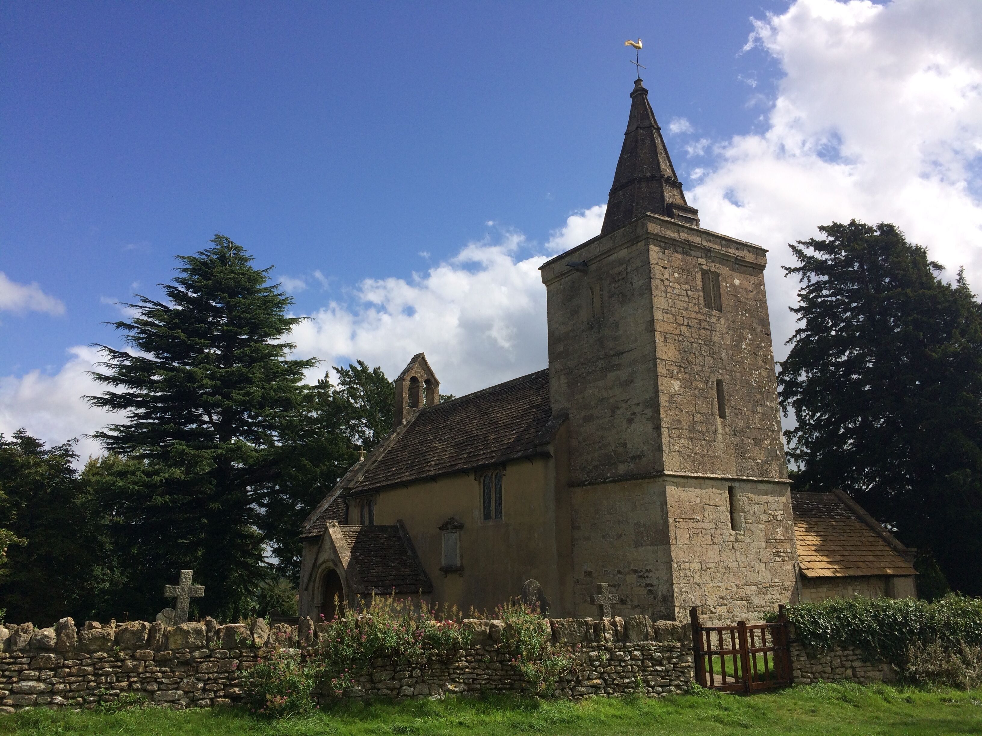 A church with a tall tower and a weather vane, surrounded by stone walls and greenery.