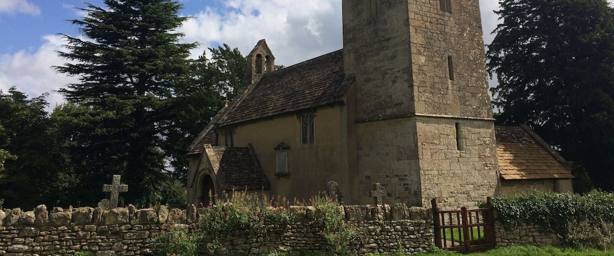 A church with a tall tower and a weather vane, surrounded by stone walls and greenery.
