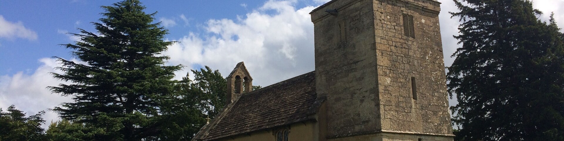 A church with a tall tower and a weather vane, surrounded by stone walls and greenery.