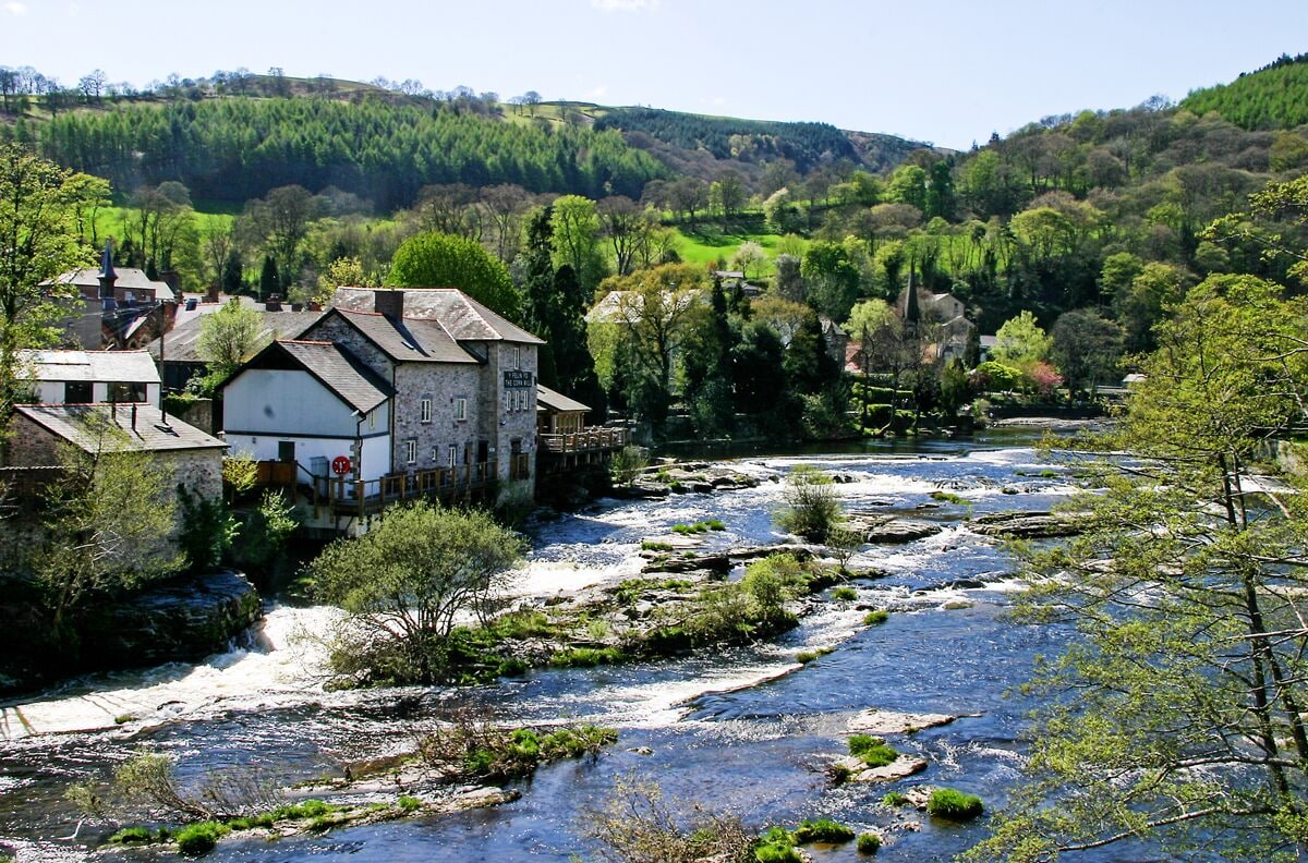 A river flowing through a village with stone buildings and lush greenery.