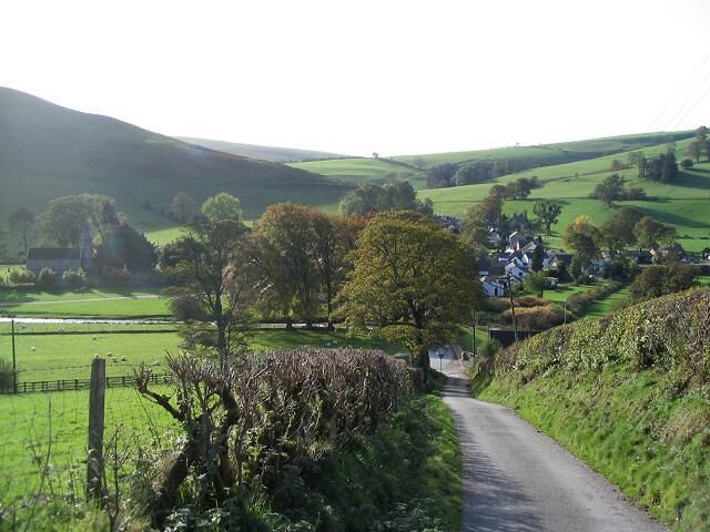 A rural landscape with a narrow road, rolling hills, and scattered houses.