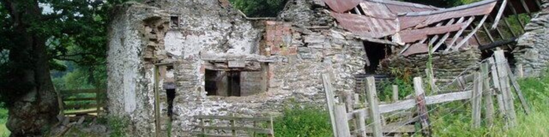 Uma estrutura de pedra deteriorada com o telhado desabado, rodeada por vegetação exuberante e um cercado de madeira.