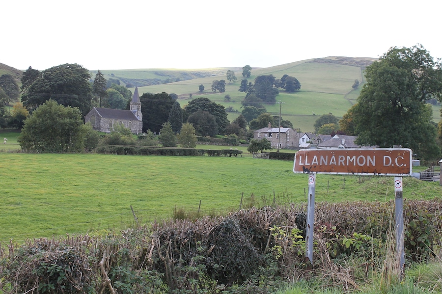Un paisaje rural con una iglesia, un cartel que dice "LLANARMON D.C." y ondulantes colinas verdes.