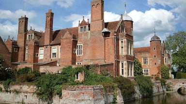 A historic brick building with multiple towers and chimneys, surrounded by greenery and a body of water.