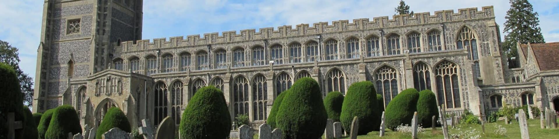 A historic stone building with a tall tower and arched windows, surrounded by a well-maintained cemetery with headstones and trimmed hedges.