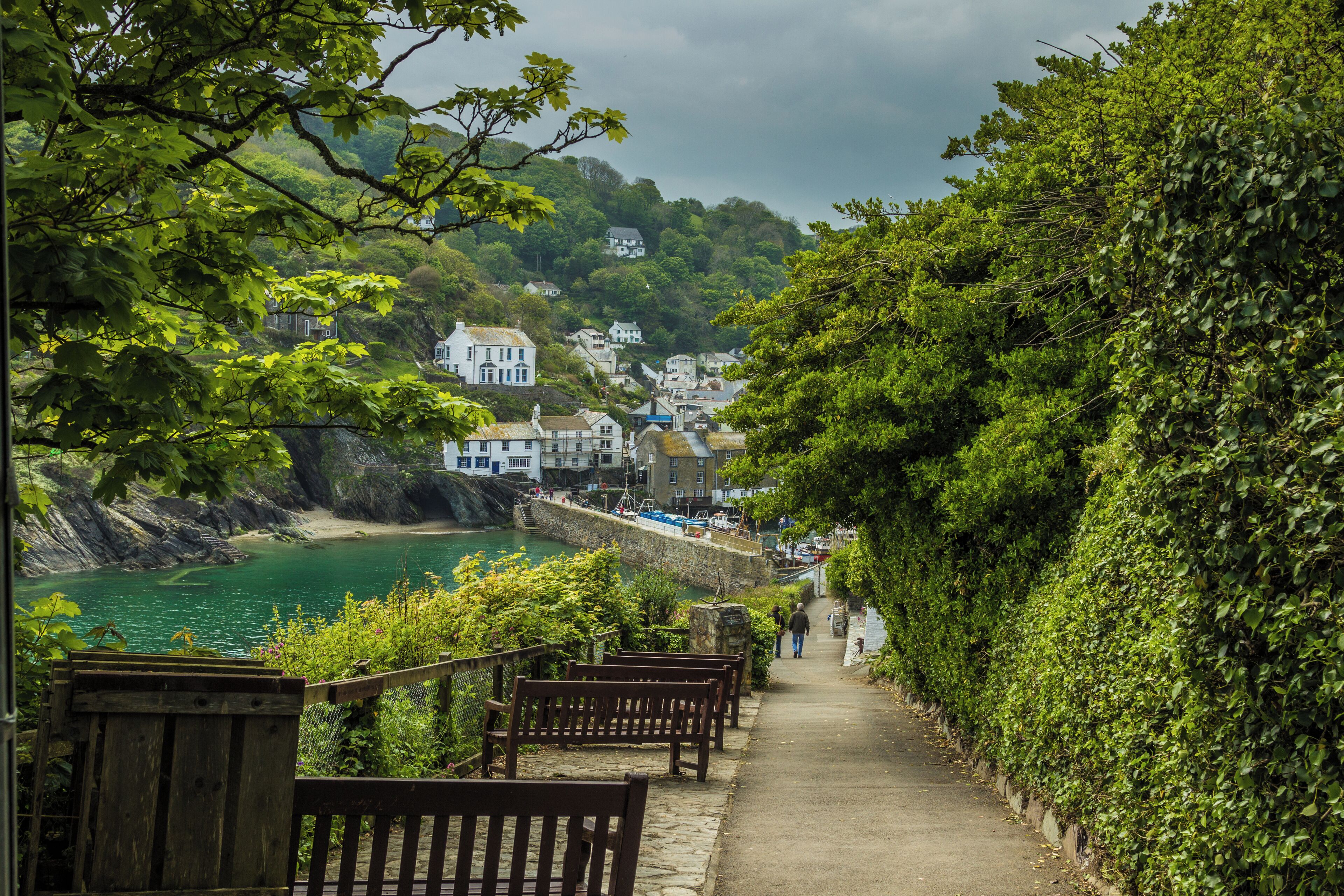 A coastal pathway with benches, a stone wall, and houses in the background.