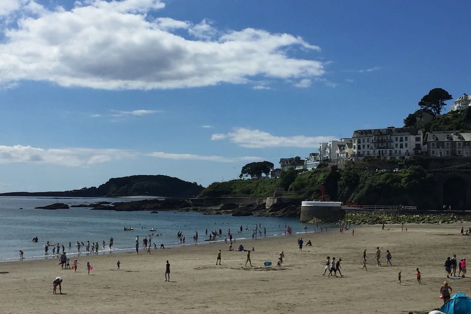 A crowded beach with people, a rocky coastline, and buildings on a hill.