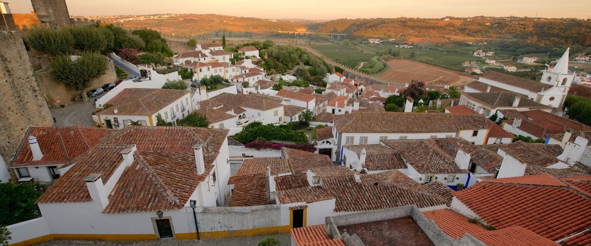 A historic town with white buildings and red-tiled roofs, surrounded by a stone wall and a tower.