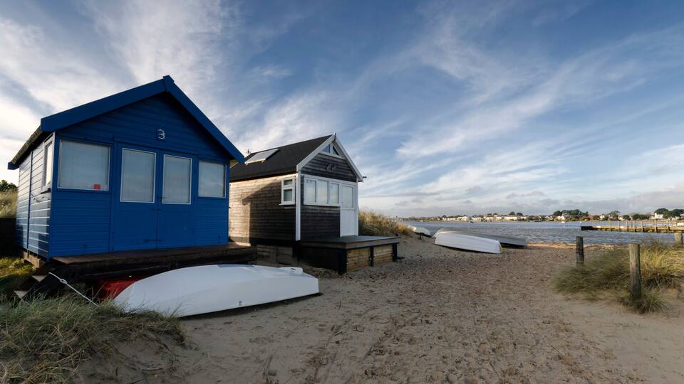 A beach with two small beach huts, one blue and one brown, and a sandy shore with a calm sea in the background.