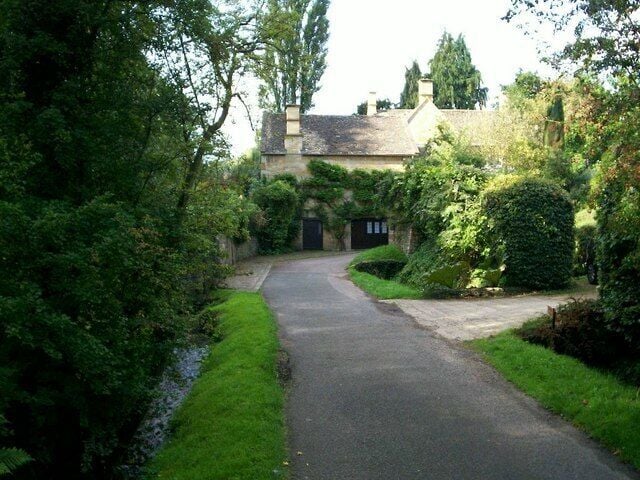 A narrow, tree-lined road leading to a stone house with a dark entrance.