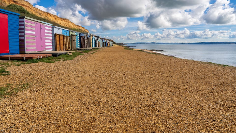 A pebbled beach with colorful beach huts and a cliff in the background.