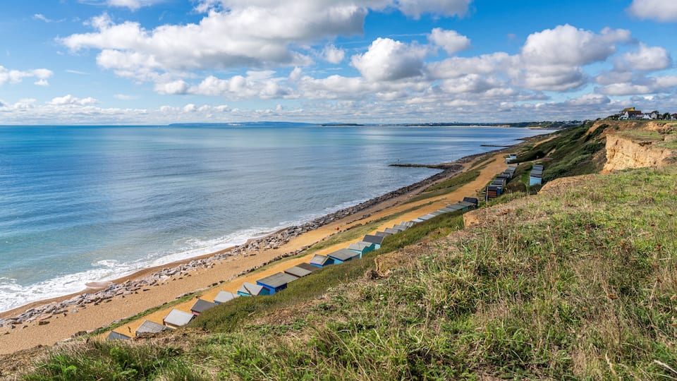 A coastal beach with a row of blue beach huts along the shore, a rocky breakwater, and a clear blue sky.