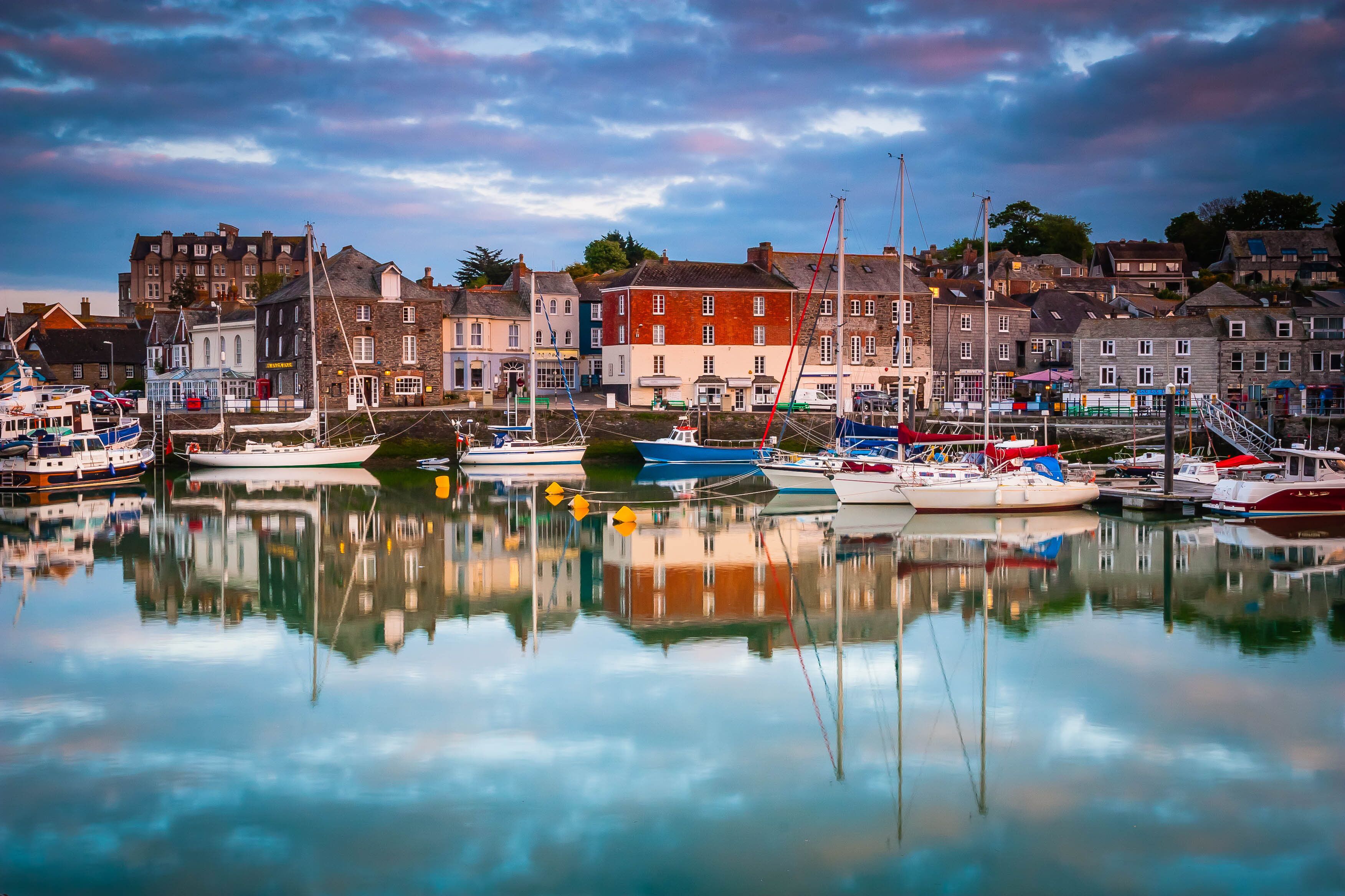 A harbor with boats docked, buildings along the waterfront, and a clear reflection in the water.