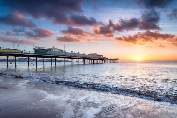 A pier extending into the water with buildings on it, a sunset in the background, and waves on the shore.