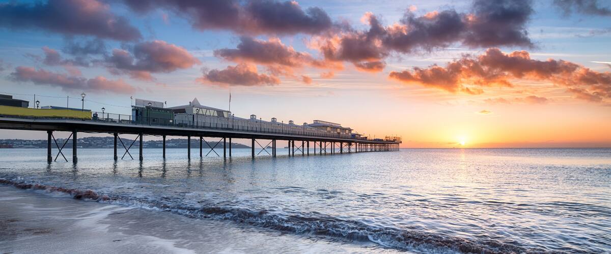 A pier extending into the water with buildings on it, a sunset in the background, and waves on the shore.