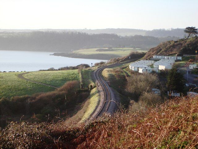 A railway track curving through a hilly landscape with a body of water in the background and a row of white buildings on the right.