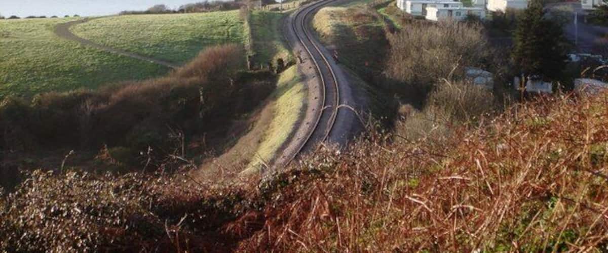 A railway track curving through a hilly landscape with a body of water in the background and a row of white buildings on the right.