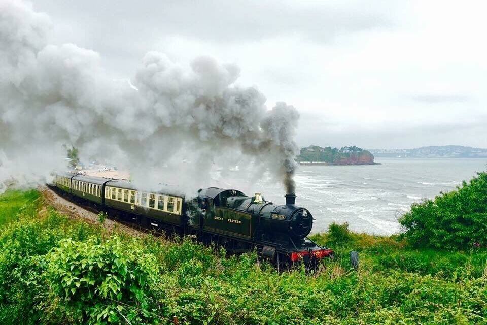 A steam locomotive with black and gold carriages traveling along a track beside a body of water.