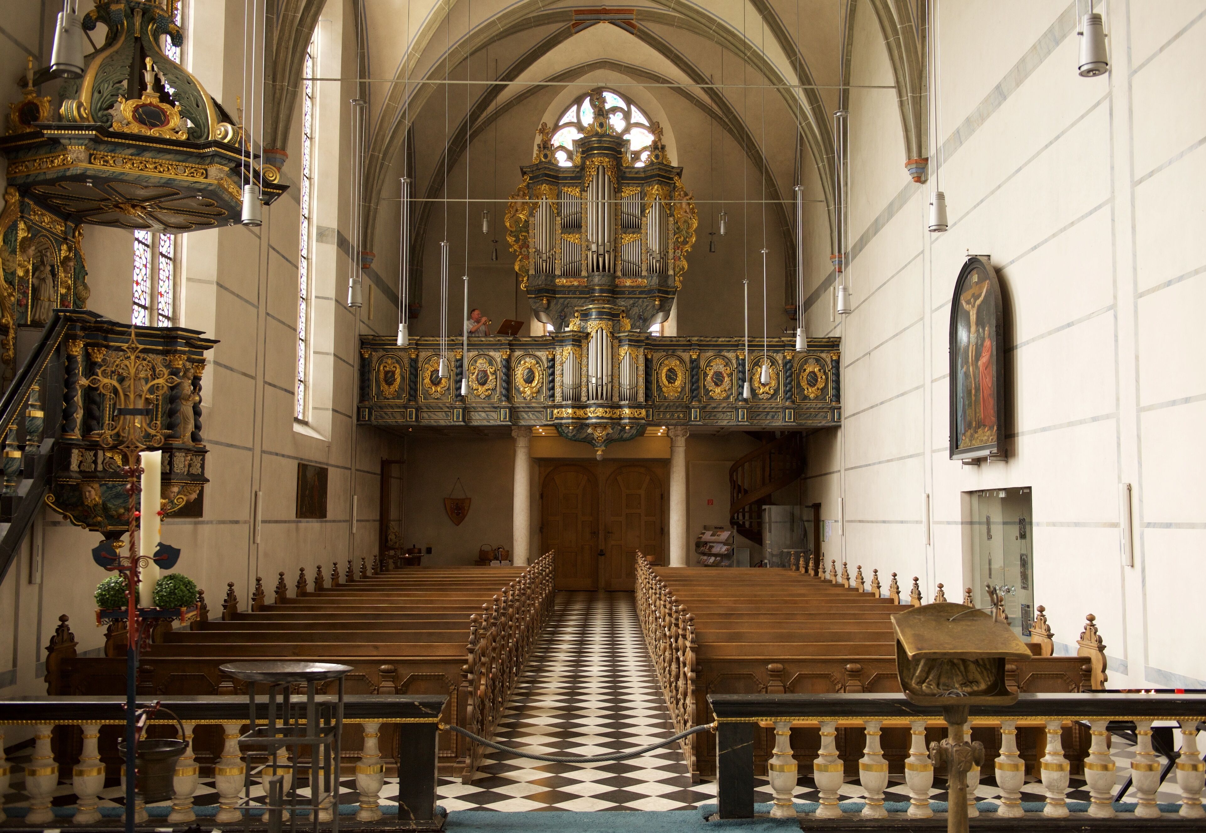 An ornate church interior with a checkered floor, pews, and a large organ.