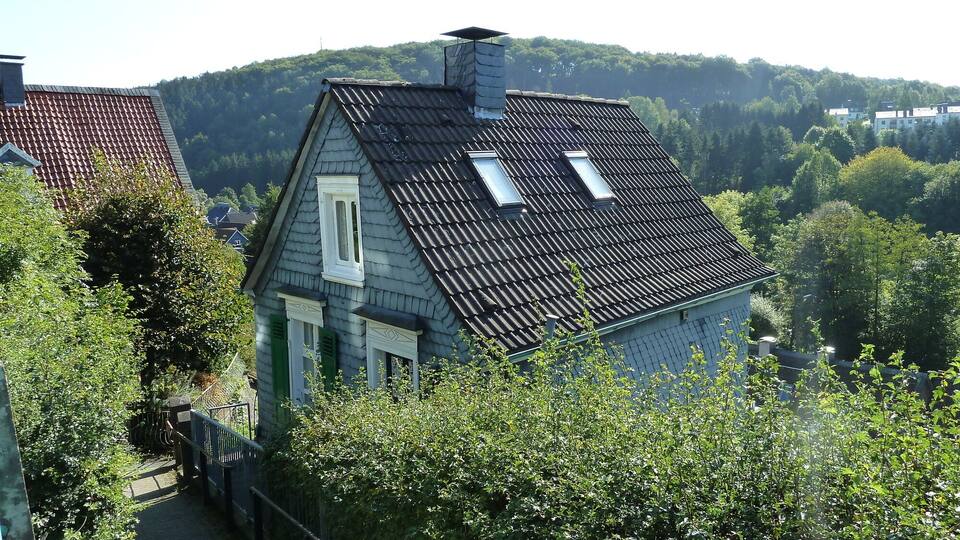 A house with a sloped roof and dormer windows, surrounded by lush greenery and hills in the background.