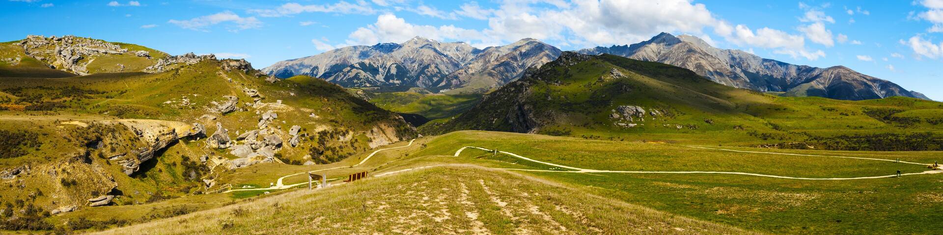 A mountainous landscape with a winding path, rocky outcrops, and distant snow-capped peaks under a clear blue sky.