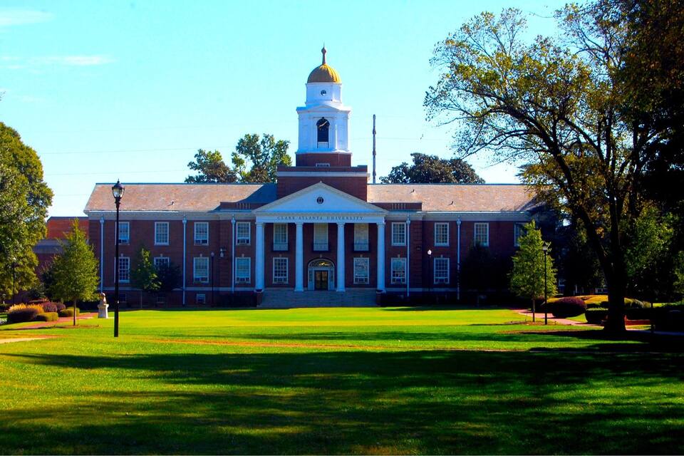 A large brick building with a central dome and a clock tower, surrounded by a well-maintained lawn and trees.