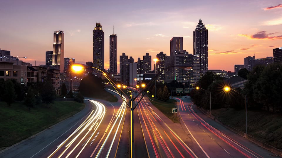 A city skyline at dusk with illuminated buildings and light trails on the roads.