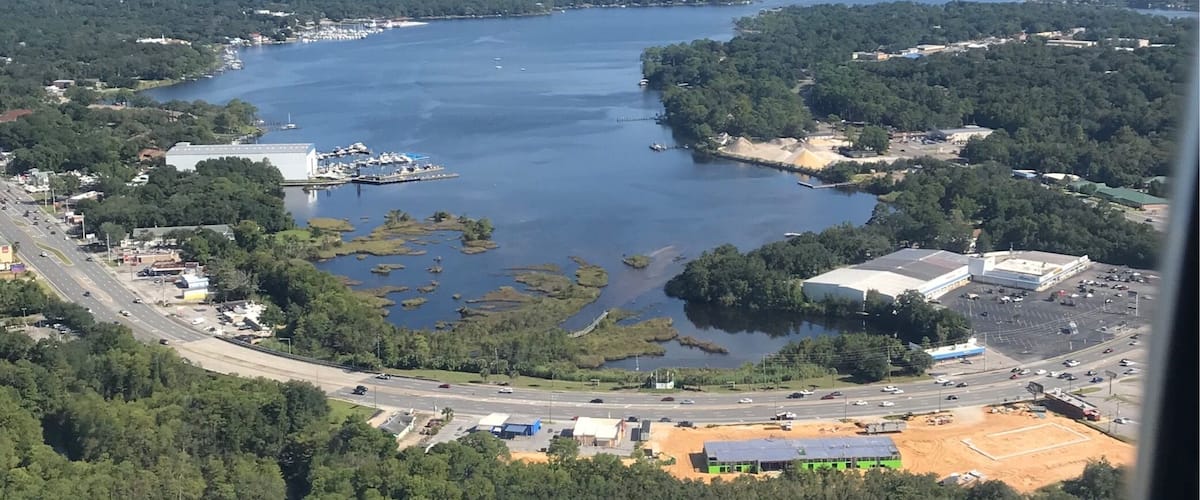 A coastal landscape with a large body of water, dense greenery, and several buildings including a large white structure and a parking lot.