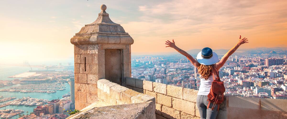 A person with arms raised, overlooking a coastal cityscape with a prominent stone tower.