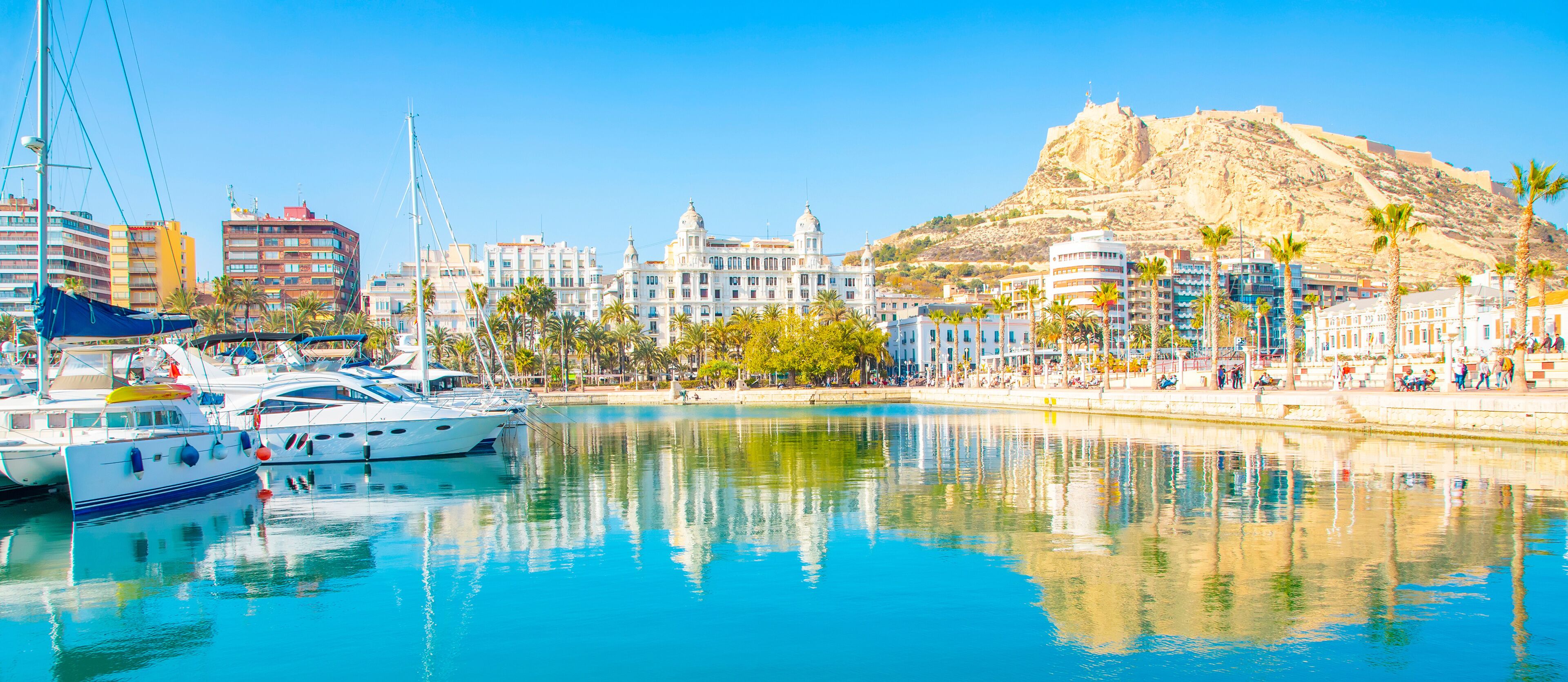 A marina with boats, a large white building with domes, a hill with a fort, and palm trees.