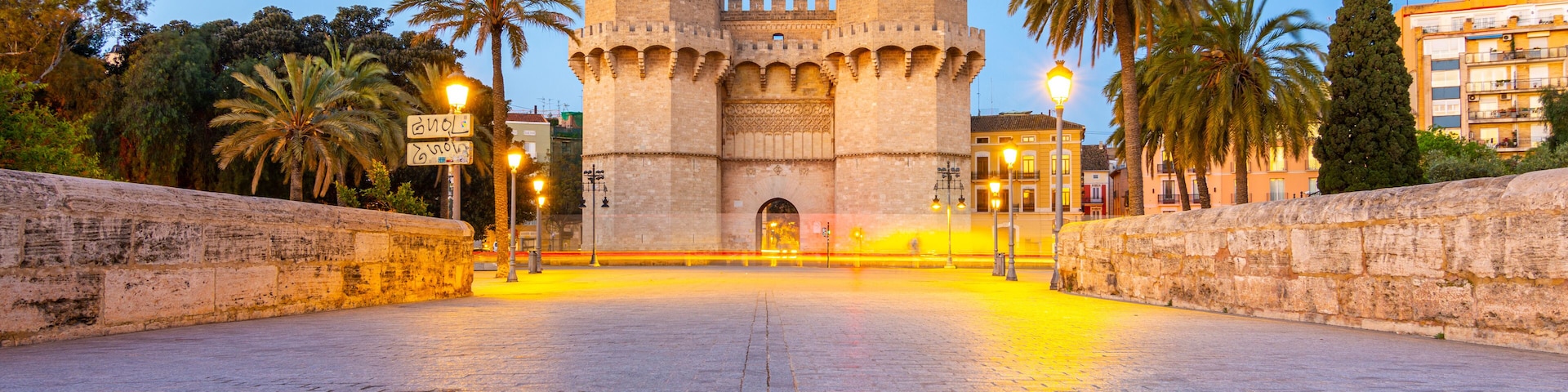 Un castillo histórico con dos torres, palmeras y un área de entrada empavedada.
