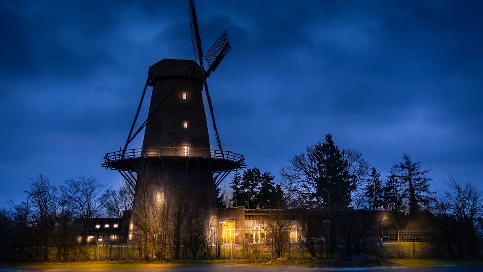 A windmill at dusk with lights on, surrounded by trees and buildings.