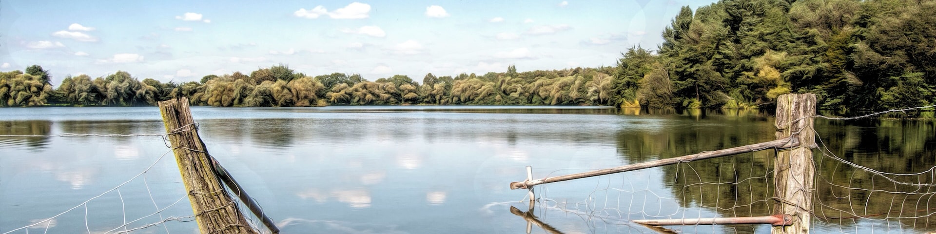 Un lago tranquilo con un poste de madera y una valla de alambre en primer plano, rodeado de árboles densos y un cielo azul despejado.