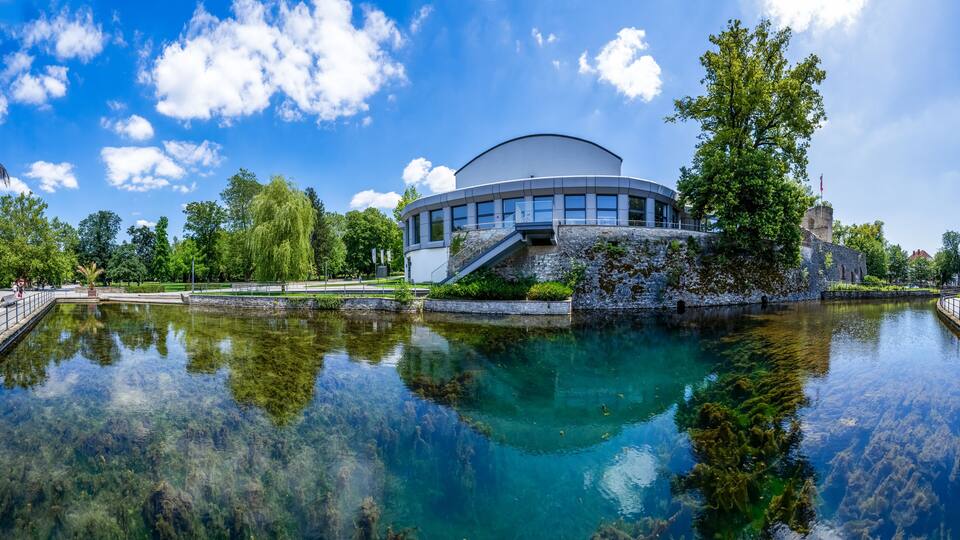 A modern building with a circular design, a clear water body reflecting the structure, and surrounding greenery.