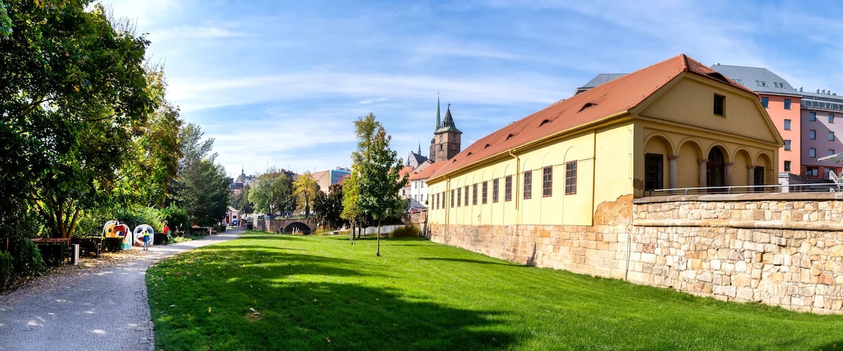 A historic building with a stone wall, a church tower, and a paved path.
