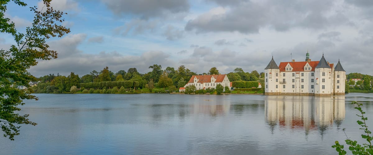 A large castle by a calm lake with surrounding greenery and other buildings.