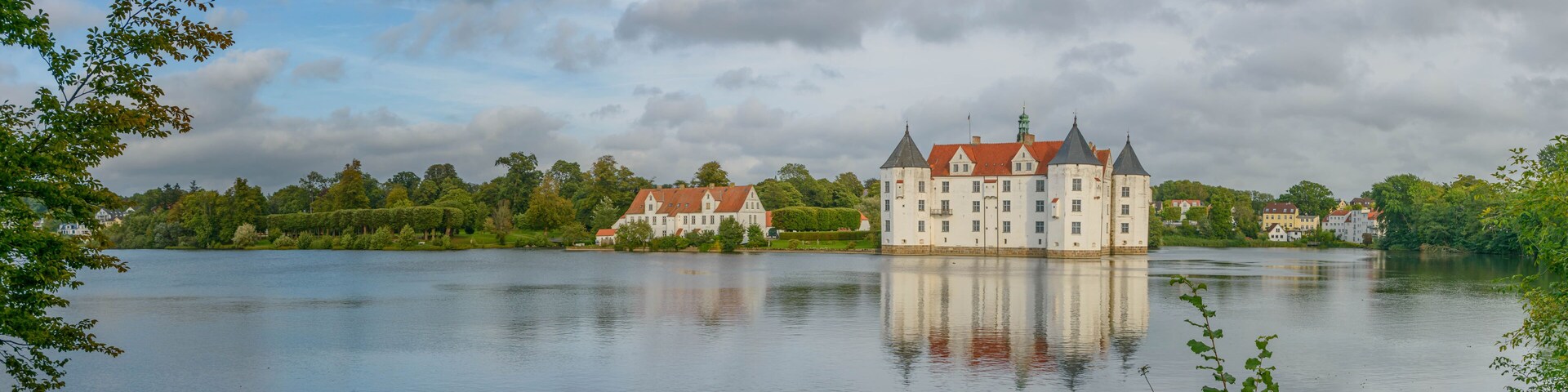 A large castle by a calm lake with surrounding greenery and other buildings.