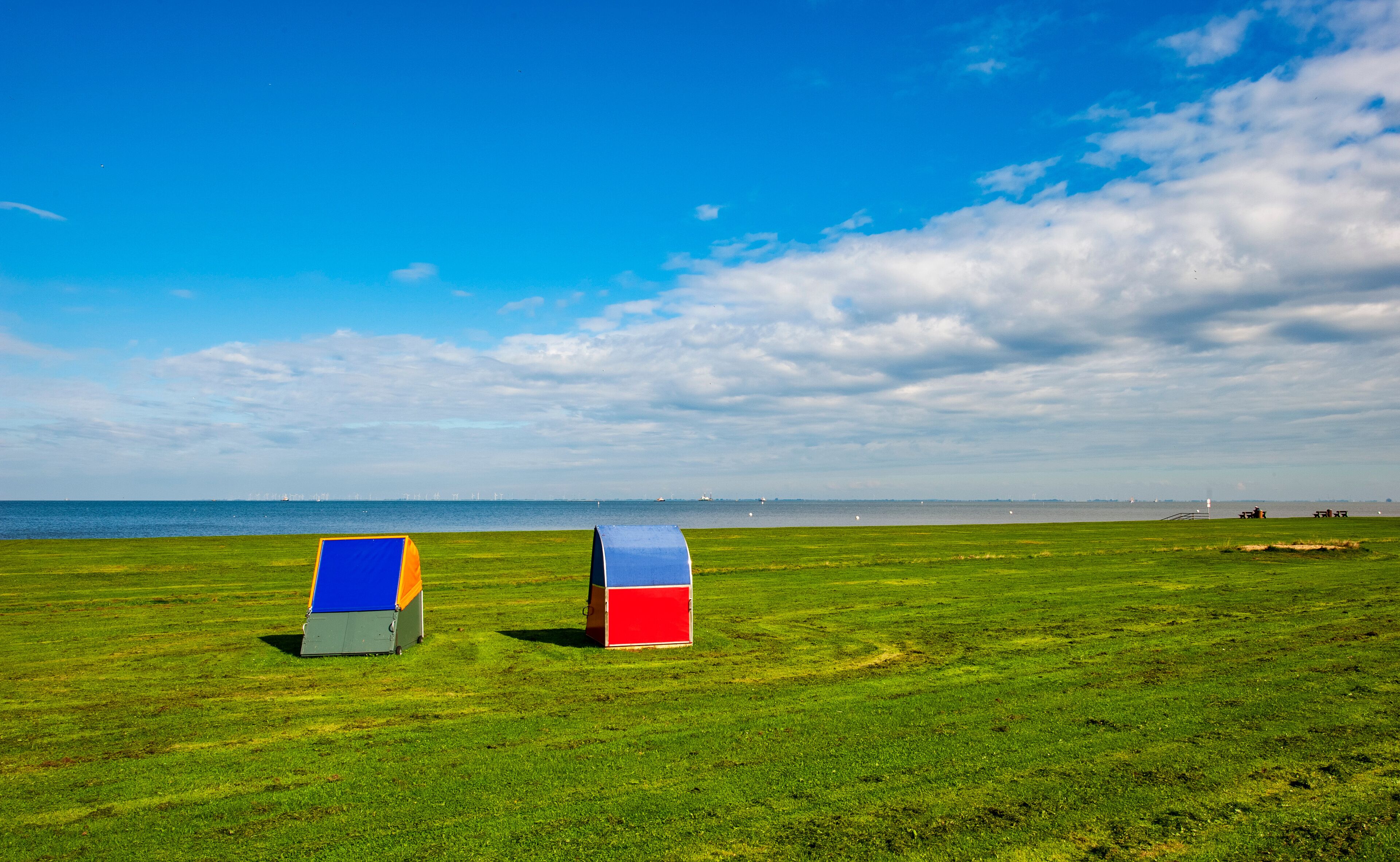 Two colorful beach umbrellas on a grassy field with a view of the sea and distant wind turbines.