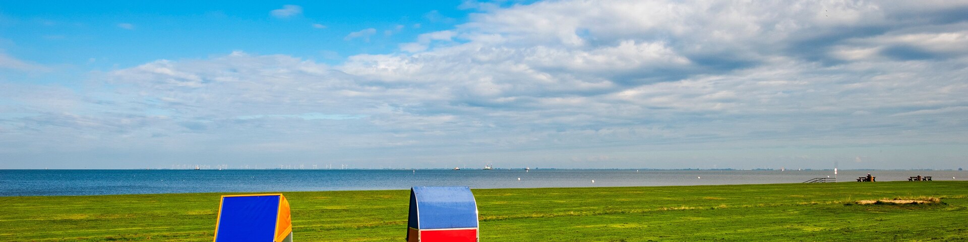 Two colorful beach umbrellas on a grassy field with a view of the sea and distant wind turbines.
