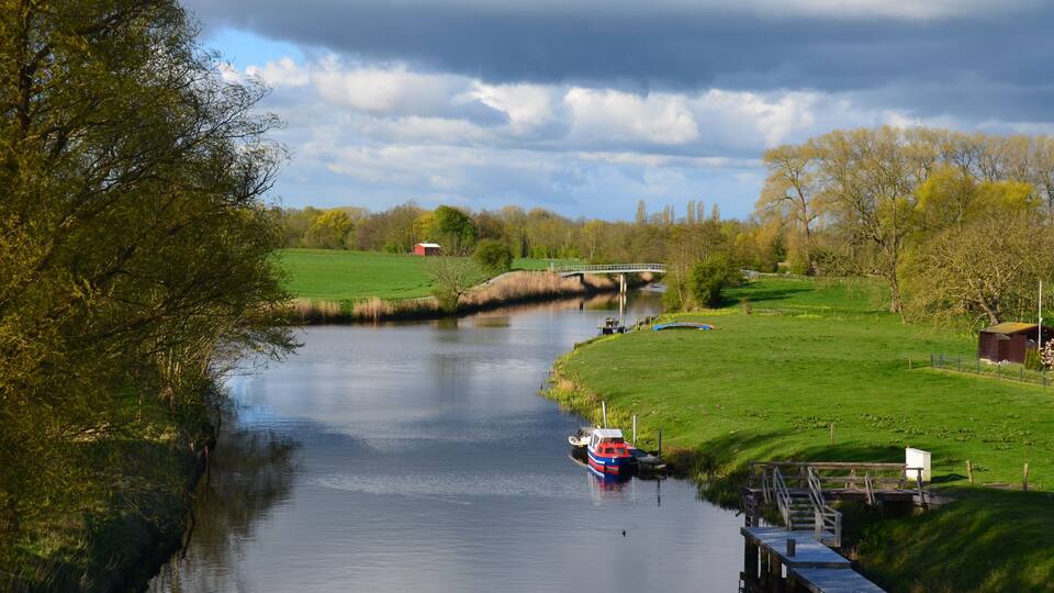 A river with a boat docked on the right, a bridge in the background, and green fields on both sides.