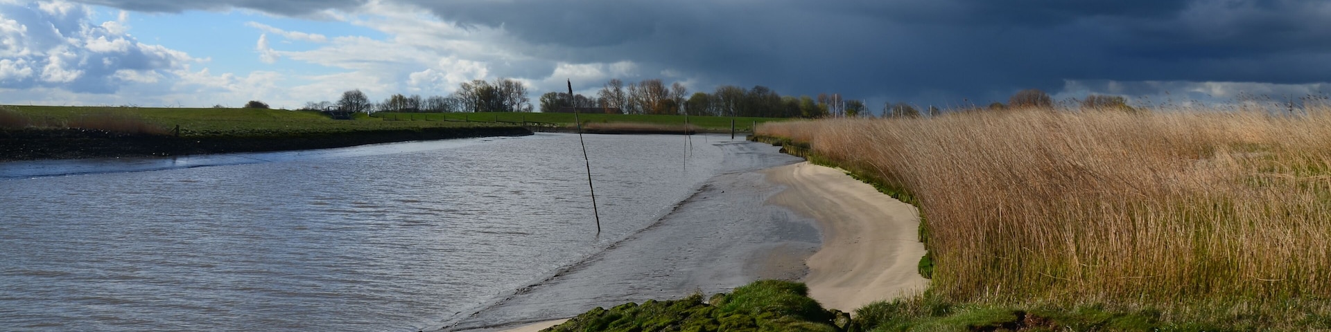 Orilla de río con playa de arena, zonas de césped y cielo nublado.