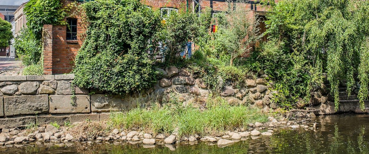 A traditional half-timbered house by a calm river with a stone wall and lush greenery.