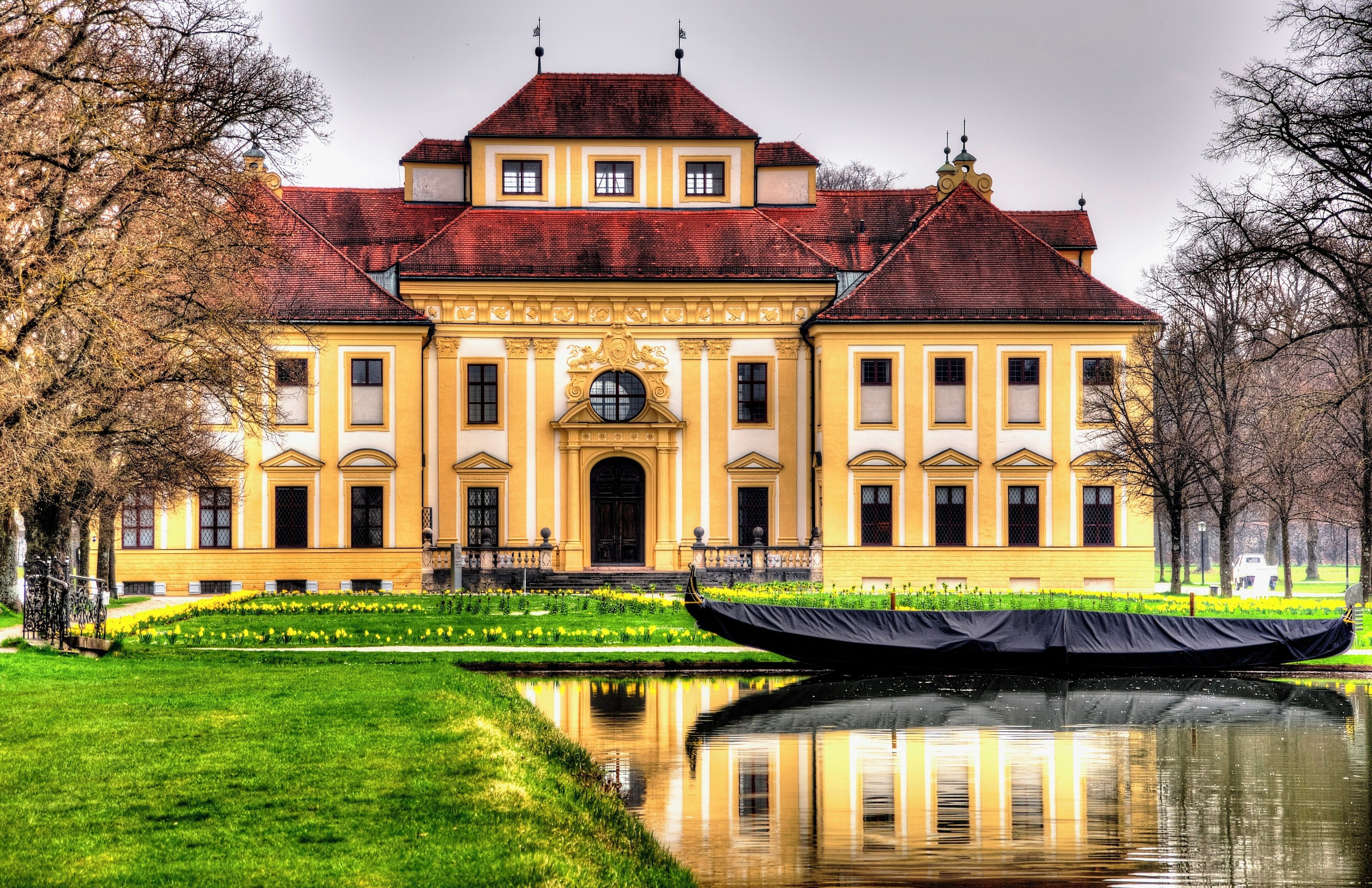 A large yellow building with a red roof, surrounded by a pond and trees.