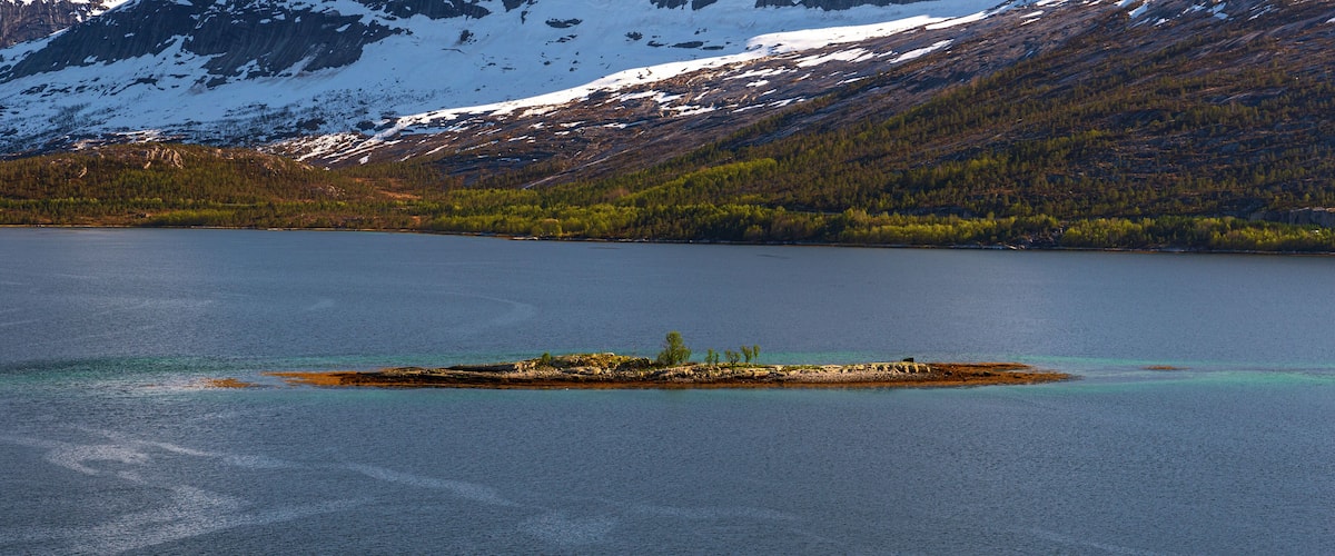 A lake with a small island in the center, surrounded by mountains with snow-capped peaks and dense forests.