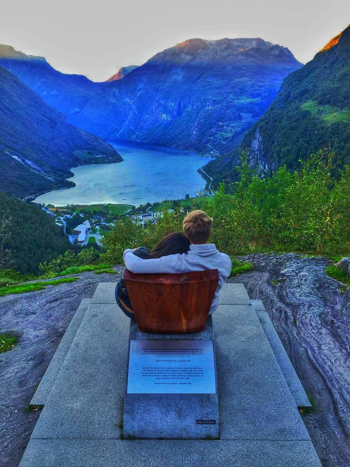 A couple sitting on a bench overlooking a valley with a lake and mountains.