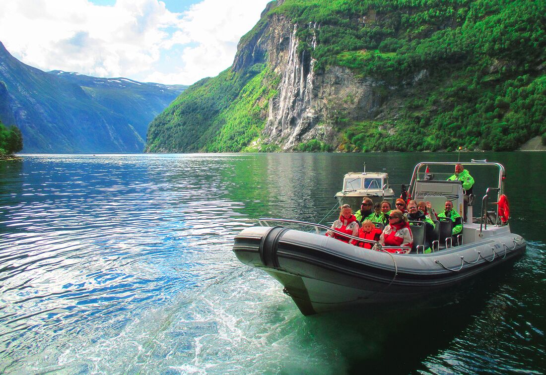 A boat with passengers on a calm lake surrounded by lush green mountains and a waterfall.