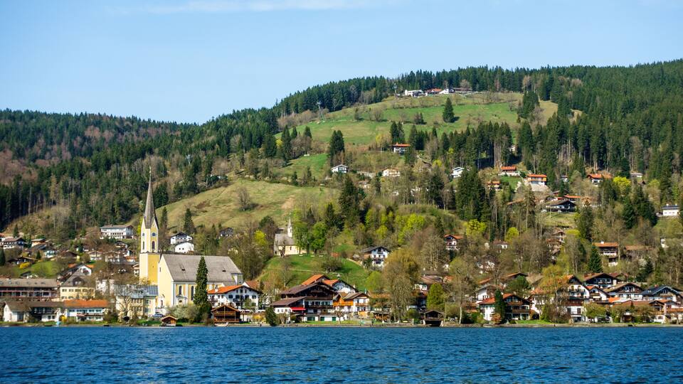 A lakeside village with houses, a church, and a hill with trees.