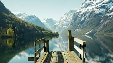 A wooden dock extending into a calm lake surrounded by snow-capped mountains and dense forest.