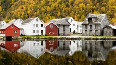 A row of wooden houses by a calm lake with autumn foliage in the background.