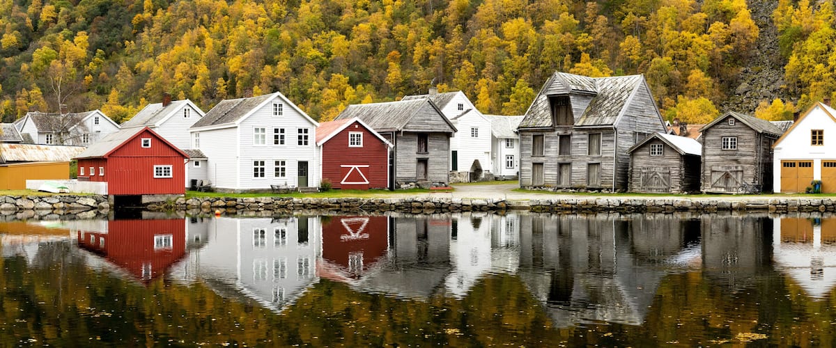 A row of wooden houses by a calm lake with autumn foliage in the background.